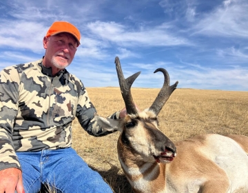 A hunter wearing camouflage and an orange cap poses with a pronghorn antelope in a golden Wyoming field under a sky streaked with soft white clouds.